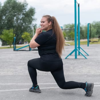 A beautiful fat girl in a black tracksuit is engaged in fitness on the sports ground. Young woman lunges outdoors on a warm summer day. Healthy lifestyle and weight loss