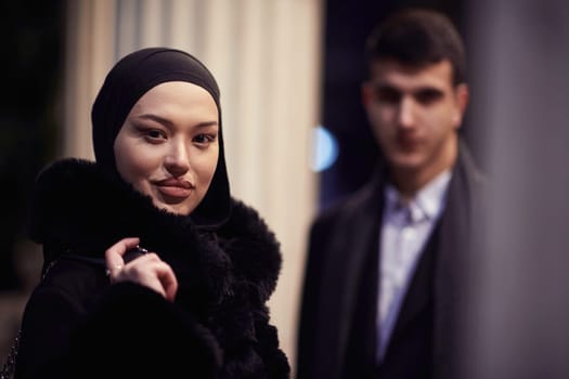 Happy multicultural business couple walking together outdoors in an urban city street at night near a jewelry shopping store window. Successful Arab businessman and European Muslim woman.