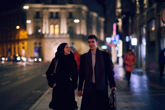 Happy multicultural business couple walking together outdoors in an urban city street at night near a jewelry shopping store window. Successful Arab businessman and European Muslim woman.