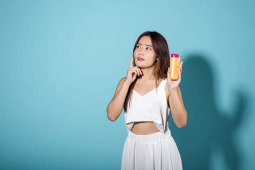 Beautiful Asian young woman holding body lotion on hand in studio shot isolated on blue background, Smiling brunette female hold cream sunblock bottle, tube of sunscreen, protect sun light and UV