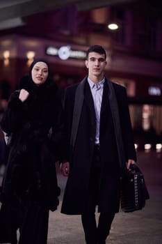 Happy multicultural business couple walking together outdoors in an urban city street at night near a jewelry shopping store window. Successful Arab businessman and European Muslim woman.