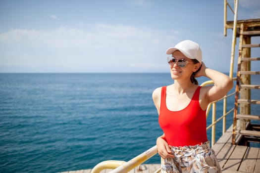 A beautiful girl in a red swimsuit with long hair, glasses and a white cap looks at the blue sea.