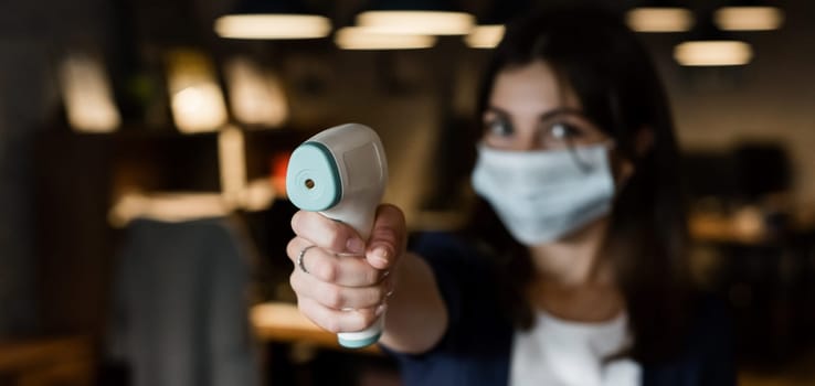 A female office worker in a mask holds a contactless electronic thermometer.