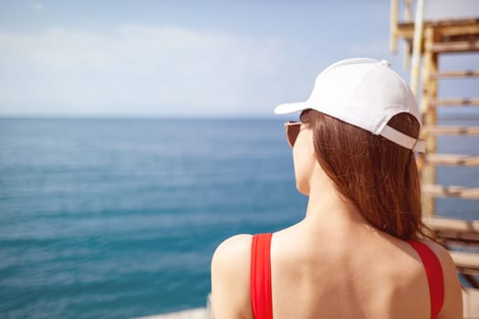 A beautiful girl in a red swimsuit with long hair, glasses and a white cap looks at the blue sea.