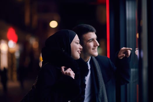 Happy multicultural business couple walking together outdoors in an urban city street at night near a jewelry shopping store window. Successful Arab businessman and European Muslim woman.