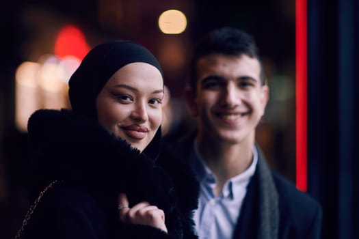 Happy multicultural business couple walking together outdoors in an urban city street at night near a jewelry shopping store window. Successful Arab businessman and European Muslim woman.