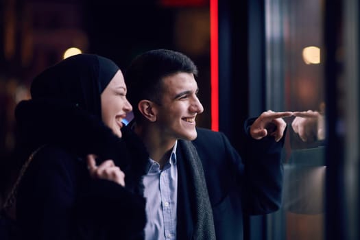 Happy multicultural business couple walking together outdoors in an urban city street at night near a jewelry shopping store window. Successful Arab businessman and European Muslim woman.