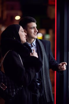 Happy multicultural business couple walking together outdoors in an urban city street at night near a jewelry shopping store window. Successful Arab businessman and European Muslim woman.