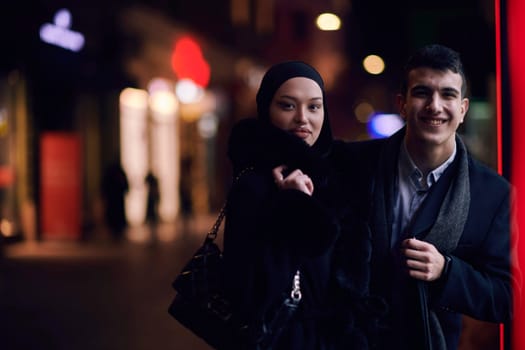 Happy multicultural business couple walking together outdoors in an urban city street at night near a jewelry shopping store window. Successful Arab businessman and European Muslim woman.