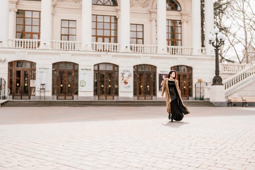 Woman street lifestyle. Image of stylish woman walking through European city on sunny day. Pretty woman with dark flowing hair, dressed in a beige raincoat and black, walks along the building