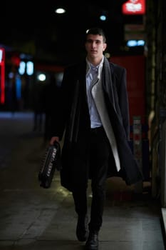 Smiling Meedle Eastern man walking down street near modern office building, freelancer businessman looking away holding mobile phone on busy city street at night