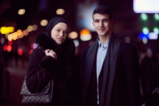 Happy multicultural business couple walking together outdoors in an urban city street at night near a jewelry shopping store window. Successful Arab businessman and European Muslim woman.