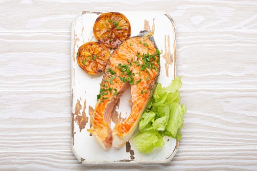 Grilled fish salmon steak and green salad with lemon served on white cutting board rustic wooden background top view, balanced diet or healthy nutrition meal with salmon and veggies.