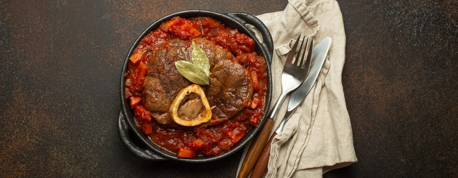 Traditional Italian dish Ossobuco all Milanese made with cut veal shank meat with vegetable tomato sauce served in black casserole pan top view on rustic brown background.