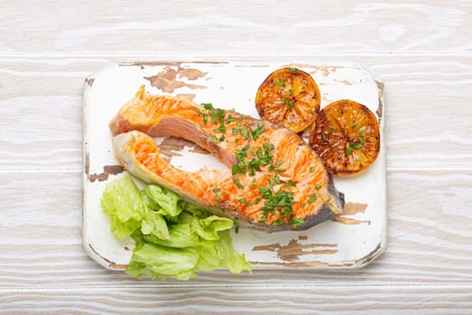 Grilled fish salmon steak and green salad with lemon served on white cutting board rustic wooden background top view, balanced diet or healthy nutrition meal with salmon and veggies.