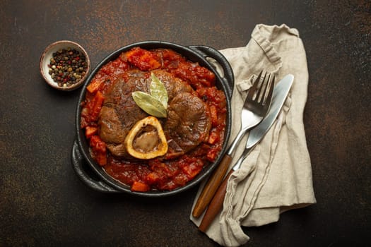 Traditional Italian dish Ossobuco all Milanese made with cut veal shank meat with vegetable tomato sauce served in black casserole pan top view on rustic brown background.