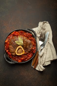 Traditional Italian dish Ossobuco all Milanese made with cut veal shank meat with vegetable tomato sauce served in black casserole pan top view on rustic brown background.