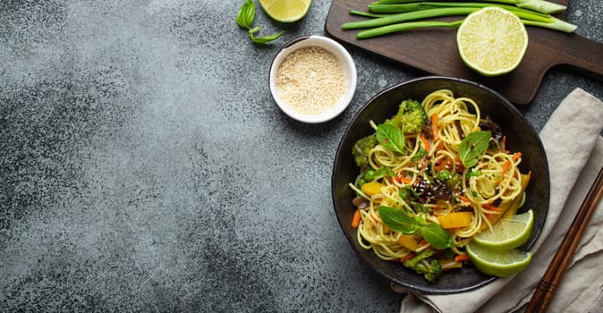 Asian vegetarian noodles with vegetables and lime in black rustic ceramic bowl, wooden chopsticks, cutting board with chopped green onion top view on stone background. Cooking noodles, copy space