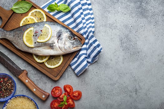 Raw fish dorado with ingredients lemon, fresh basil, cut cherry tomatoes, uncooked rice on wooden cutting board with knife on rustic stone background top view, cooking healthy fish dorado. Copy space