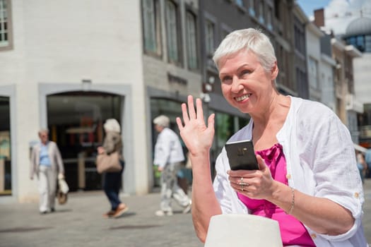 a sixty year old caucasian woman in a hat and stylish summer clothes waves to a passerby and smiles and holds a smartphone in his other hand, sits on a bench in the city, High quality photo