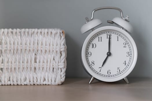 Close-up of a round white alarm clock on a table in the bedroom. The hands on the clock show seven o'clock in the morning, time to get up. Retro alarm clock on the table, vintage tone. space for text