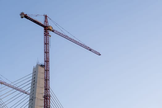 Crane and bridge construction against a blue sky background. Builders work on large construction sites, and there are many cranes working. There are a lot of cables on the bridge