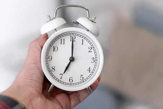 Round white alarm clock in a man's hand close-up. The hands on the clock show seven o'clock in the morning, time to get up. Retro alarm clock on the table, vintage tone. space for text