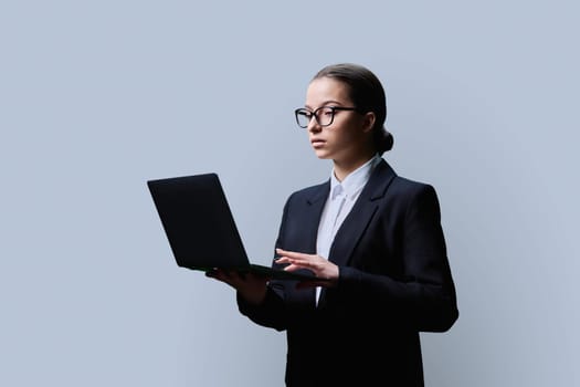 Serious teenage female student in formal business style using computer laptop, on grey studio background. Digital technologies, education, virtual educational services, online learning, youth concept