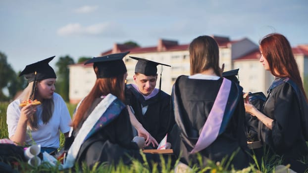 Graduates in black suits eating pizza in a city meadow