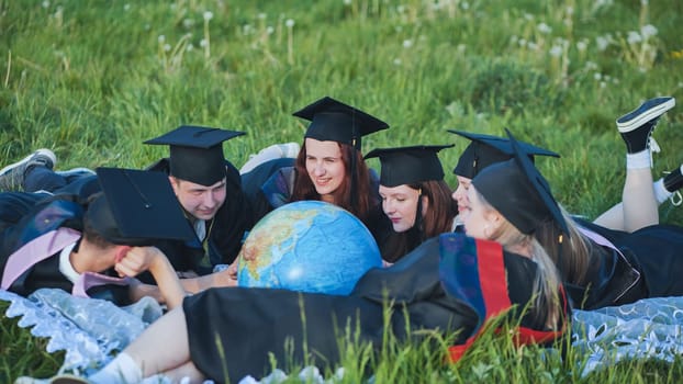 Graduate students in black robes study a globe on the grass