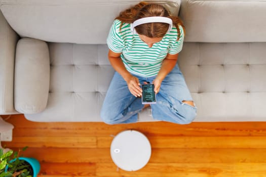 A woman wearing headphones sits on a comfortable couch, relaxing and listening to music from her smartphone while a robotic vacuum cleaner does the cleaning