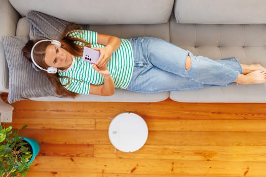 A woman wearing headphones sits on a comfortable couch, relaxing and listening to music from her smartphone while a robotic vacuum cleaner does the cleaning