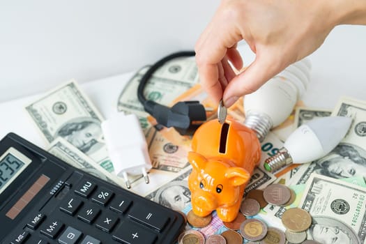A woman's hand puts a coin in an orange piggy bank in the form of a pig, saving money. Calculator, euro and dollar banknotes, coins. Tax time, bill payment, calculator for counting