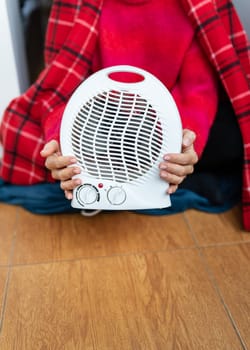 A woman in a plaid and a warm sweater warms herself at home near an electric fan heater, holds in her hands, close-up