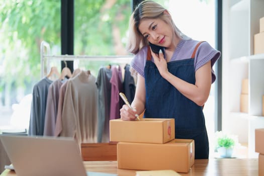 Starting small business entrepreneur of independent Asian female online seller talking on the phone with a customer and packing products for delivery to the customer. and SME delivery concept.