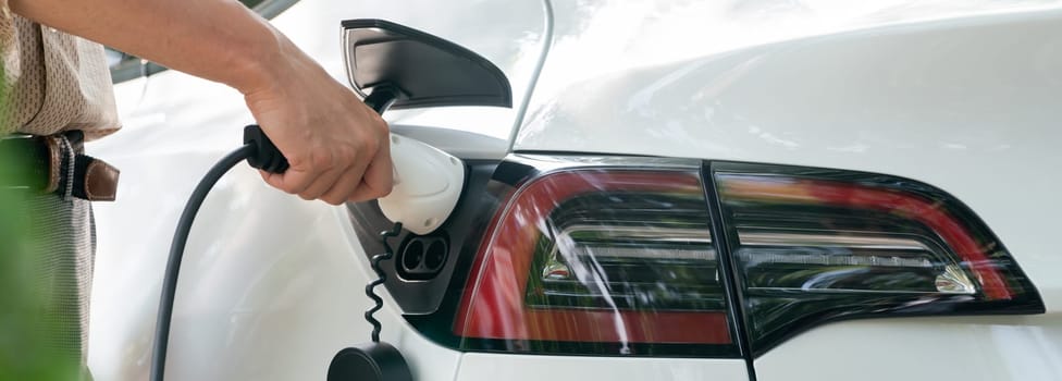 Young man recharge electric car's battery from charging station in outdoor green city park. Rechargeable EV car for sustainable environmental friendly urban travel. Panorama Expedient