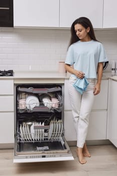 Young happy housewife woman standing with blue towel and using modern dishwasher for wash dishes at white modern kitchen.