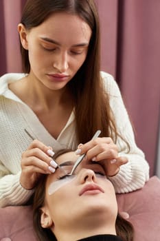 professional beautician with tweezers makes eyelash extension for beautiful woman in beauty salon.