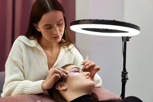 beautician with tweezers makes eyelash extension for young woman in beauty salon.