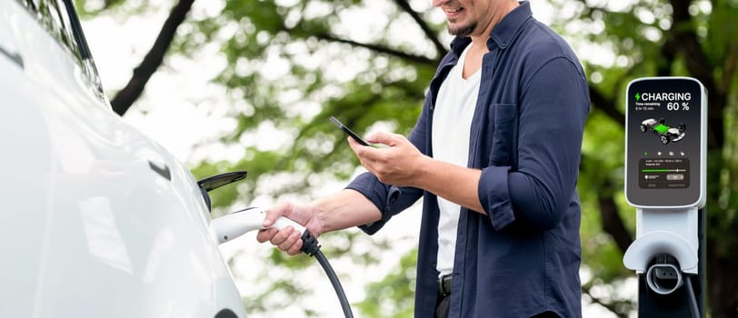 Man using smartphone online banking application to pay for electric car battery charging from EV charging station during vacation road trip at national park or summer forest. Panorama Exalt