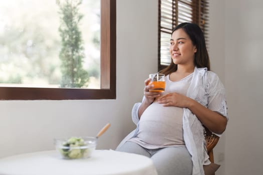 A happy pregnant woman sit, relax, drink water and eats a healthy salad on the dining room table in the living room..