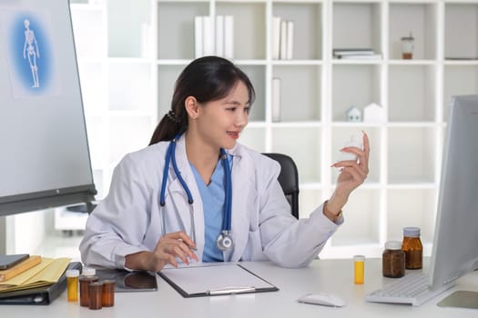 Asian female doctor holding pill bottle in hand, writing prescription, sitting at prescription table, pharmaceutical concept..