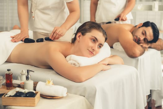 Hot stone massage at spa salon in luxury resort with day light serenity ambient, blissful couple customer enjoying spa basalt stone massage glide over body with soothing warmth. Quiescent