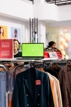 Vertical photo of open laptop with chroma key green screen standing on clothing rack during Black Friday sales in retail store. Inventory management softwares for apparel retail