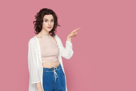 Portrait of shocked surprised woman with curly hair in casual style outfit pointing finger aside, showing advertisement area, copy space for promotion. Indoor studio shot isolated on pink background.