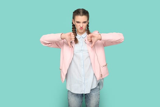 Portrait of displeased unhappy teenager girl with braids wearing pink jacket standing with thumbs down, showing dislike gesture to camera. Indoor studio shot isolated on green background.