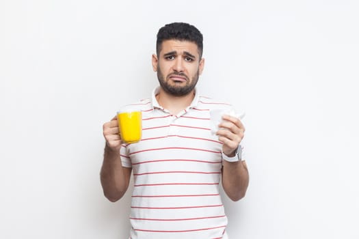 Portrait of sad unhappy unhealthy bearded man wearing striped t-shirt standing with yellow cup, looking at camera, feels bad, frowning face. Indoor studio shot isolated on gray background.