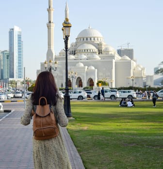 A woman in a long dress with a backpack walks along the Al Majaz embankment, Lake Khaled, Sharjah emirate. Rear view of a woman walking in the park