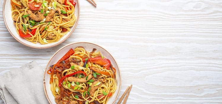 Two bowls with Chow Mein or Lo Mein, traditional Chinese stir fry noodles with meat and vegetables, served with chopsticks top view on rustic white wooden background table, space for text.