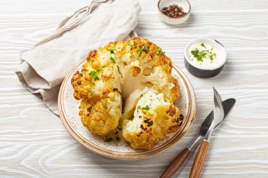 Whole roasted head of cauliflower with olive oil on plate on white wooden rustic table top view with cutlery. Vegetarian dish..